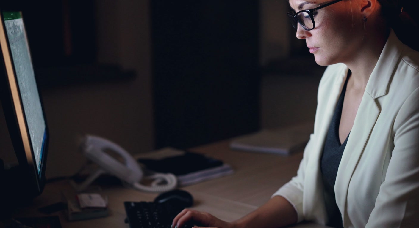 A woman working alone late at night in a dark office, lit by the glow of a computer screen