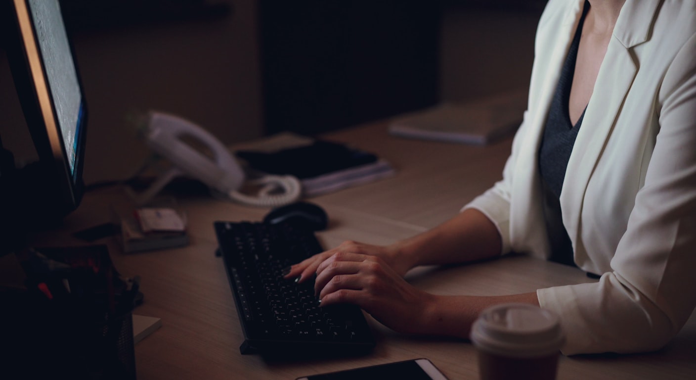 A woman working alone at a dim office desk late at night, hands poised over a keyboard